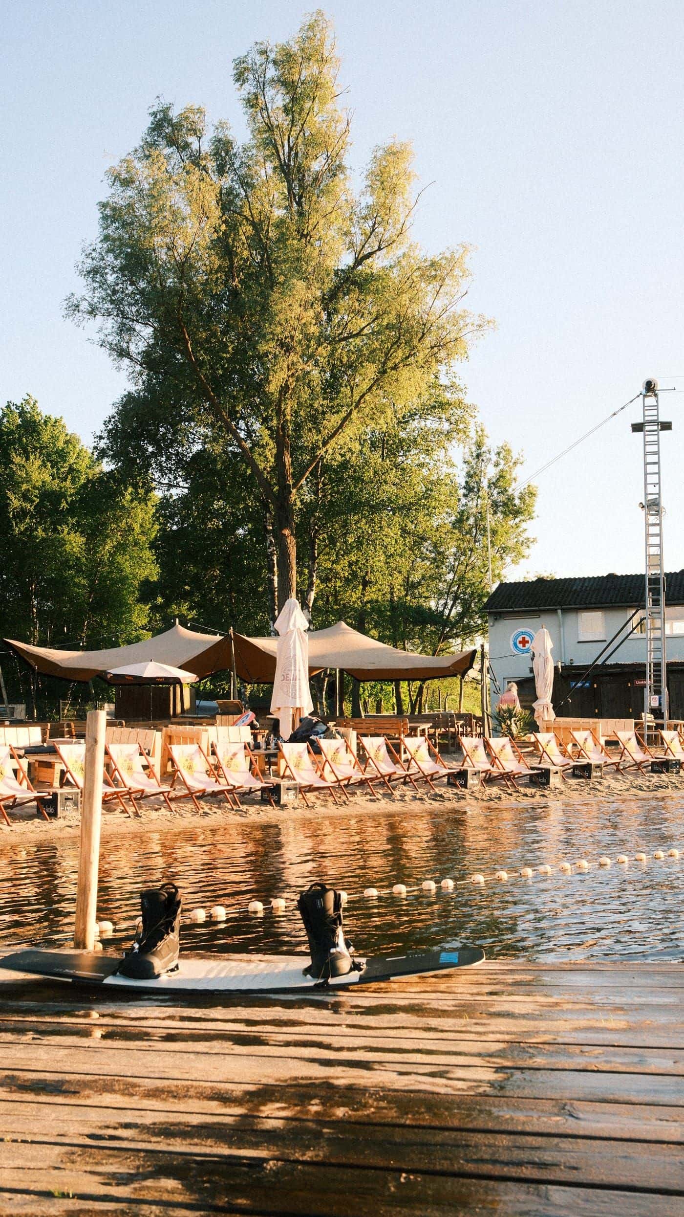 Wakeboard mit schwarzen Bindungen liegt auf dem Steg, dahinter die Liegestuhlreihe des Beachclubs und ein Mast der Seilbahn im Abendlicht