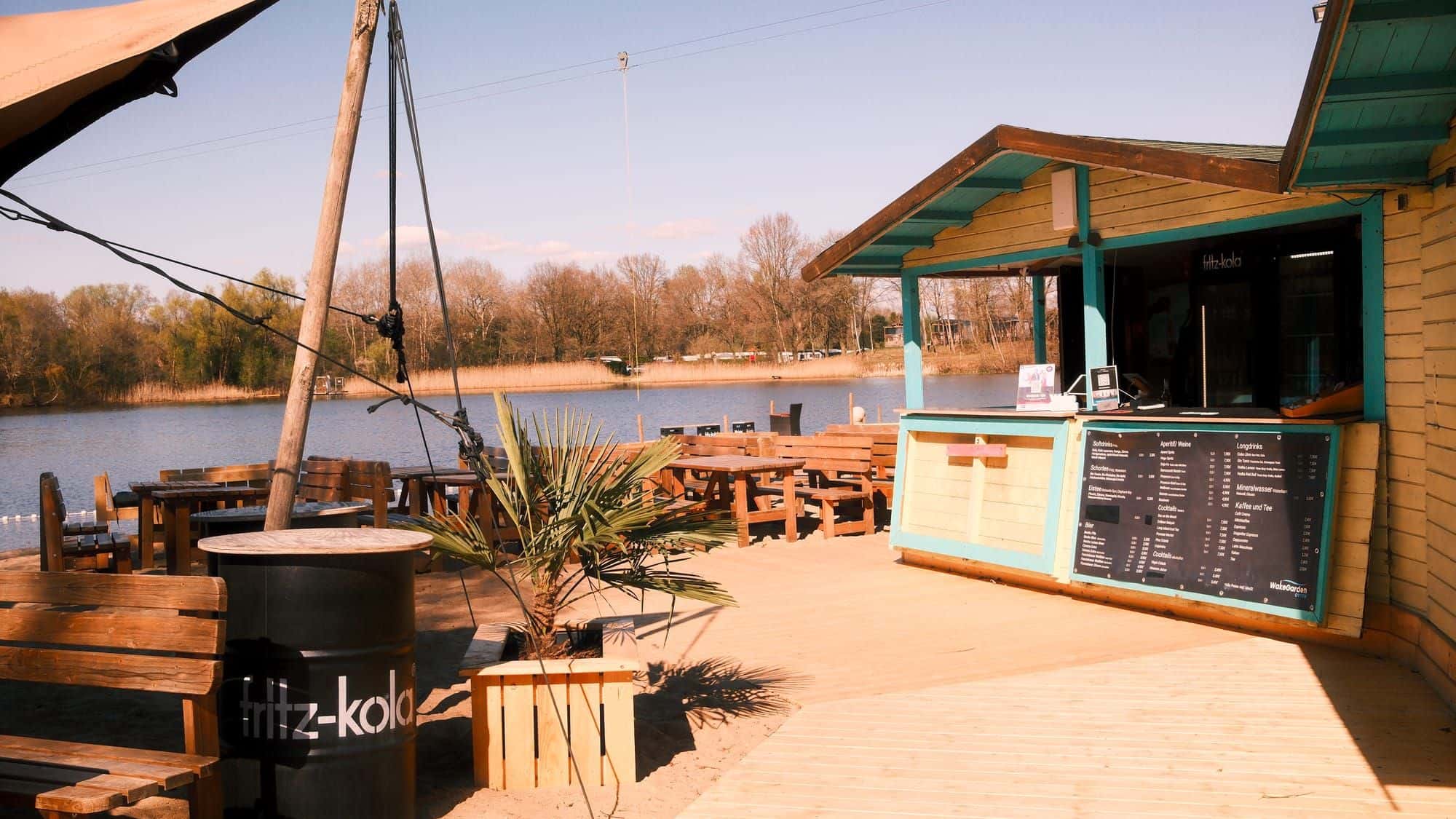 Strandbar mit türkisem Dach und Speisekarte, davor eine Palme, Fritz-Kola-Fass und Holztische am See