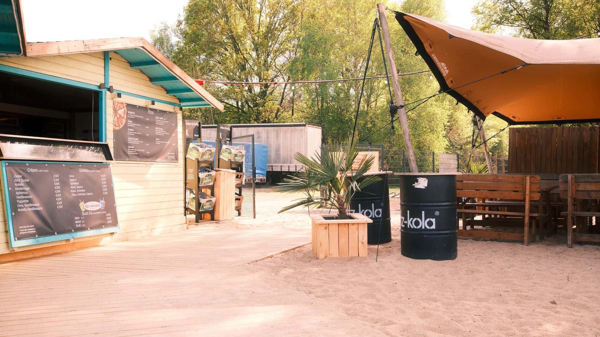 Crêpes-Hütte mit Menütafel, Palme und Fritz-Kola-Fässern auf dem Holzsteg im Sand