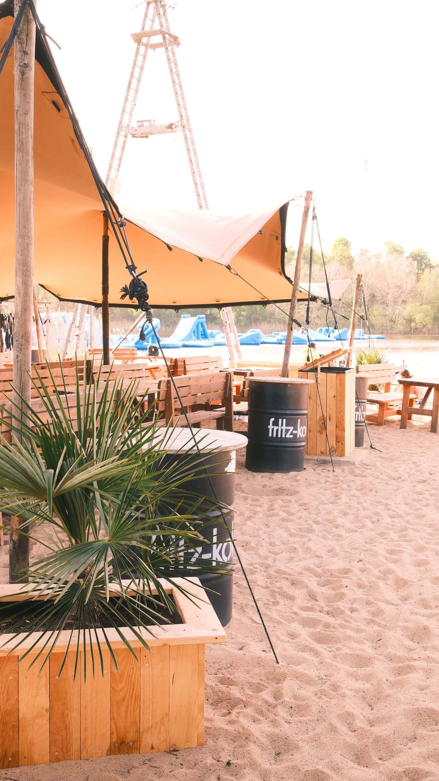 Lounge im Sand mit Palme im Holzkasten, schwarzen Fritz-Kola-Fässern und Bänken, dahinter der Aquapark