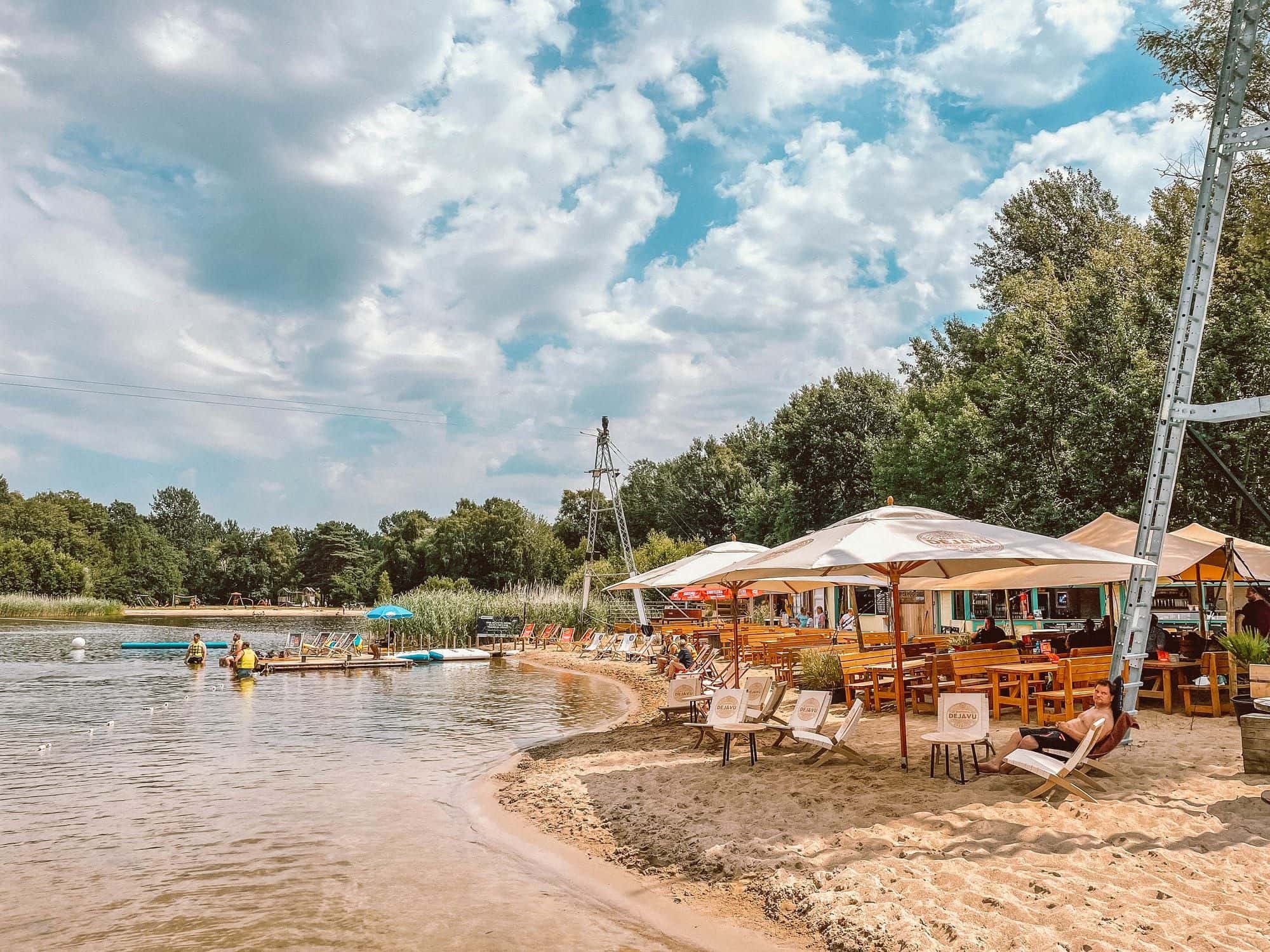 Sandstrand am Oyter See: eine Frau steht im Wasser, am Ufer Liegestühle, Sonnenschirme und der Mast der Seilbahn