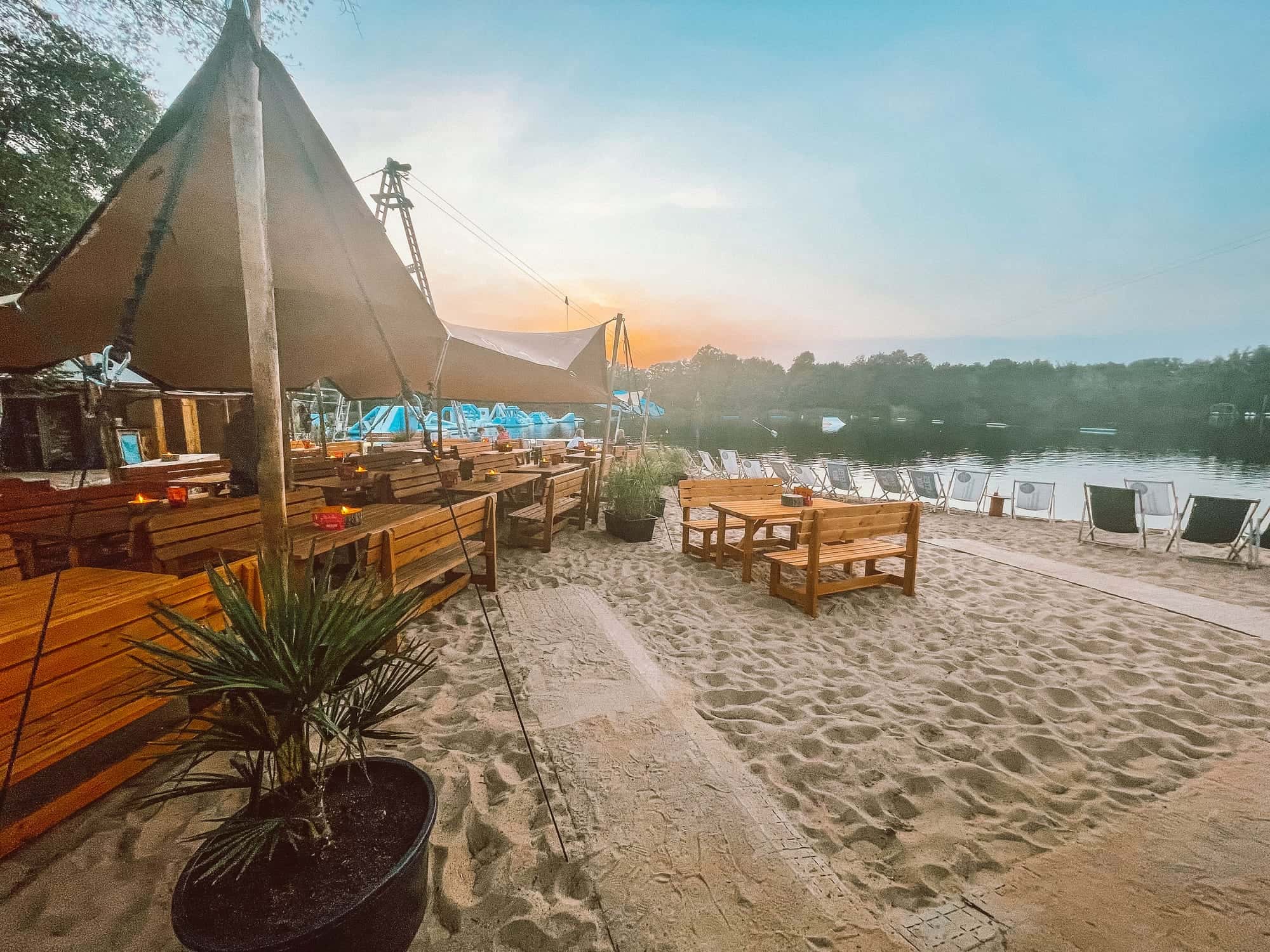 Beachclub bei Sonnenuntergang: Sonnensegel, Holzbänke und Liegestühle im Sand, der See im Abenddunst