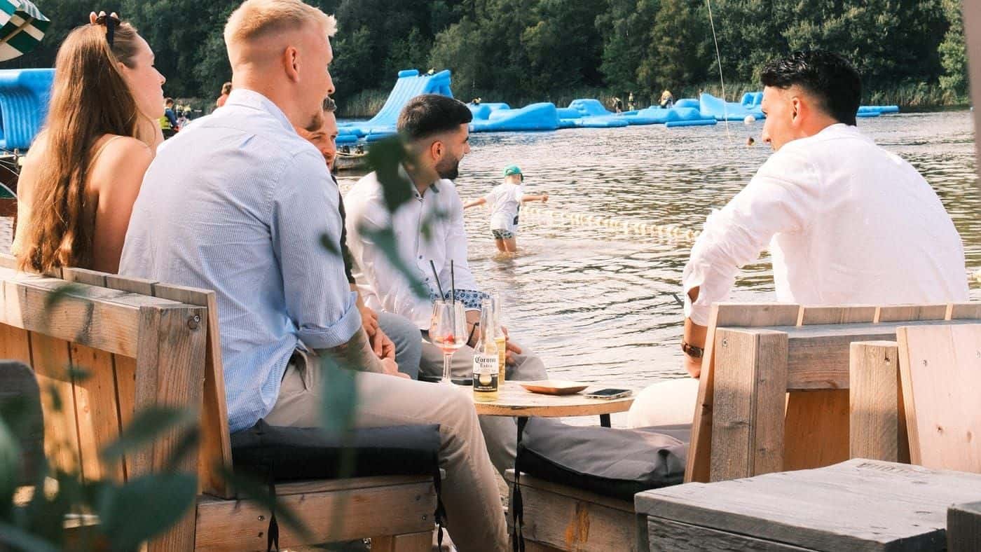 Festlich gekleidete Gäste sitzen an einem Holztisch im Sand, hinter ihnen der blaue Aquapark auf dem See