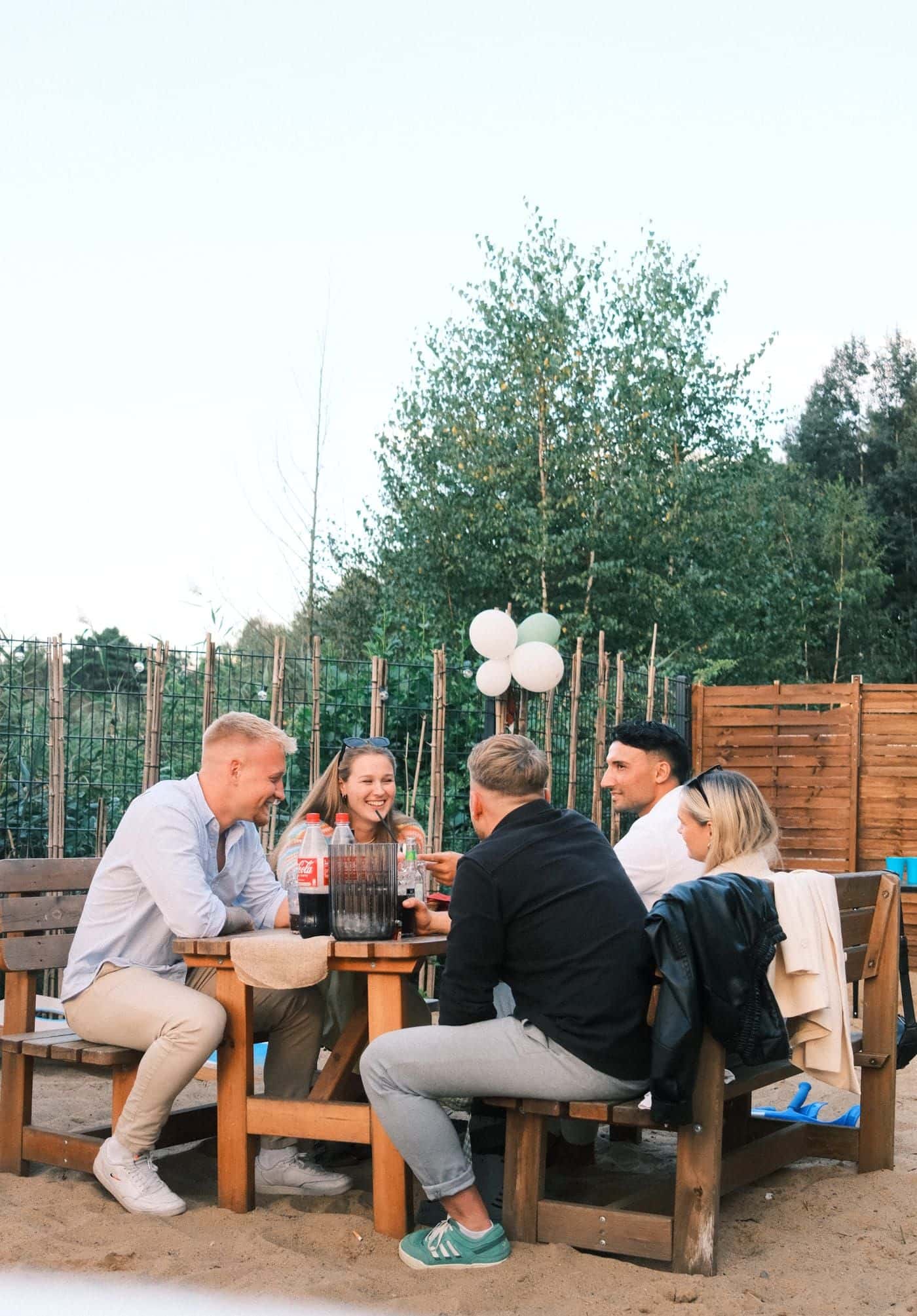 Fünf Freunde stoßen an einem Holztisch im Sand an, hinter ihnen weiße und grüne Luftballons