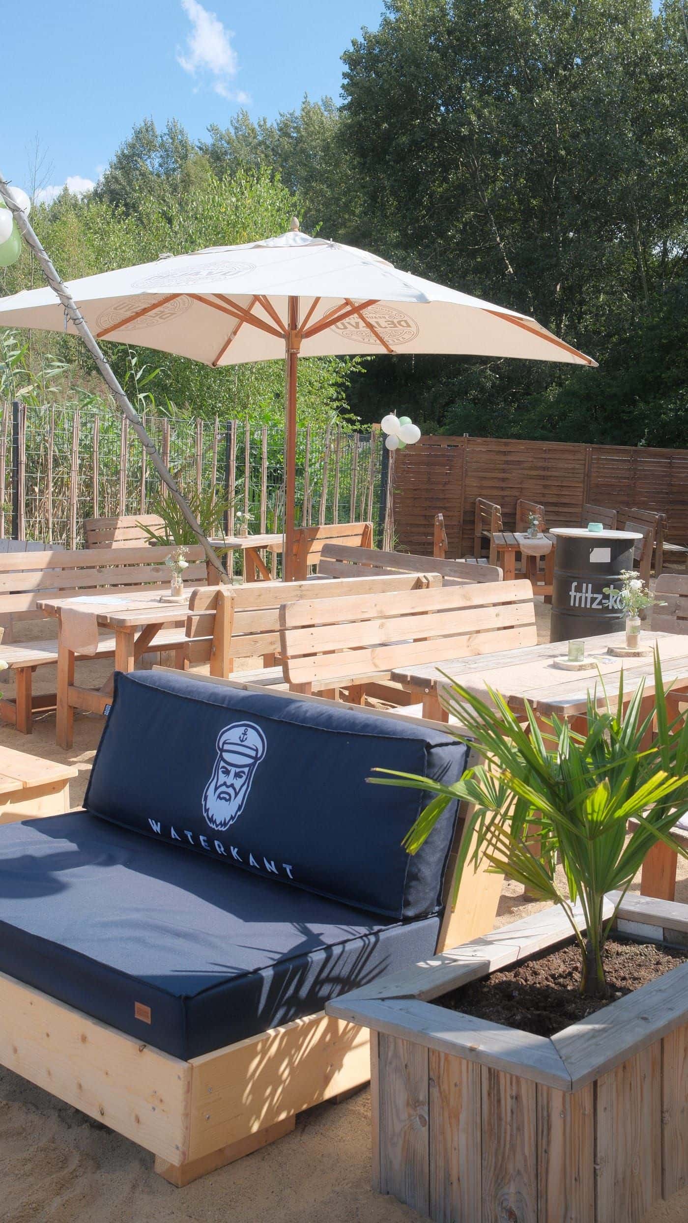 Dunkelblaues Lounge-Sofa und ein Sonnenschirm im Sand, dazwischen Tische und eine Palme