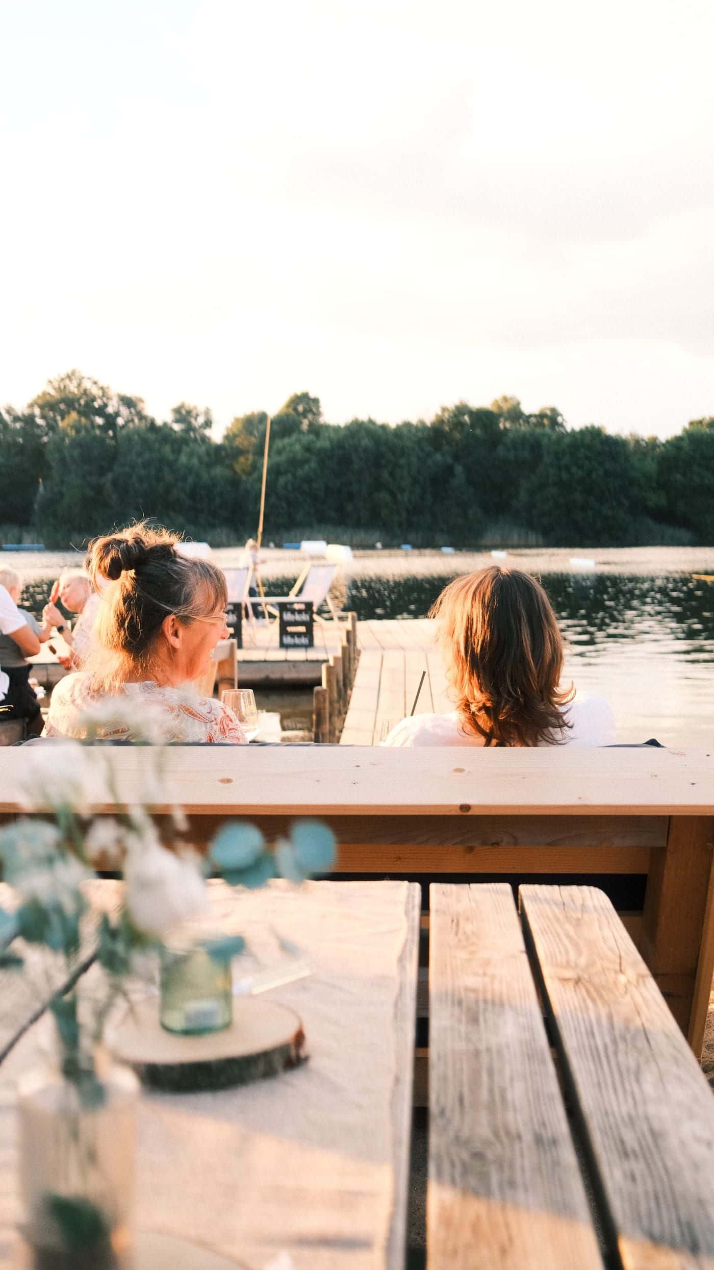 Zwei Frauen sitzen im Abendlicht mit dem Rücken zur Kamera am Steg und schauen auf den See