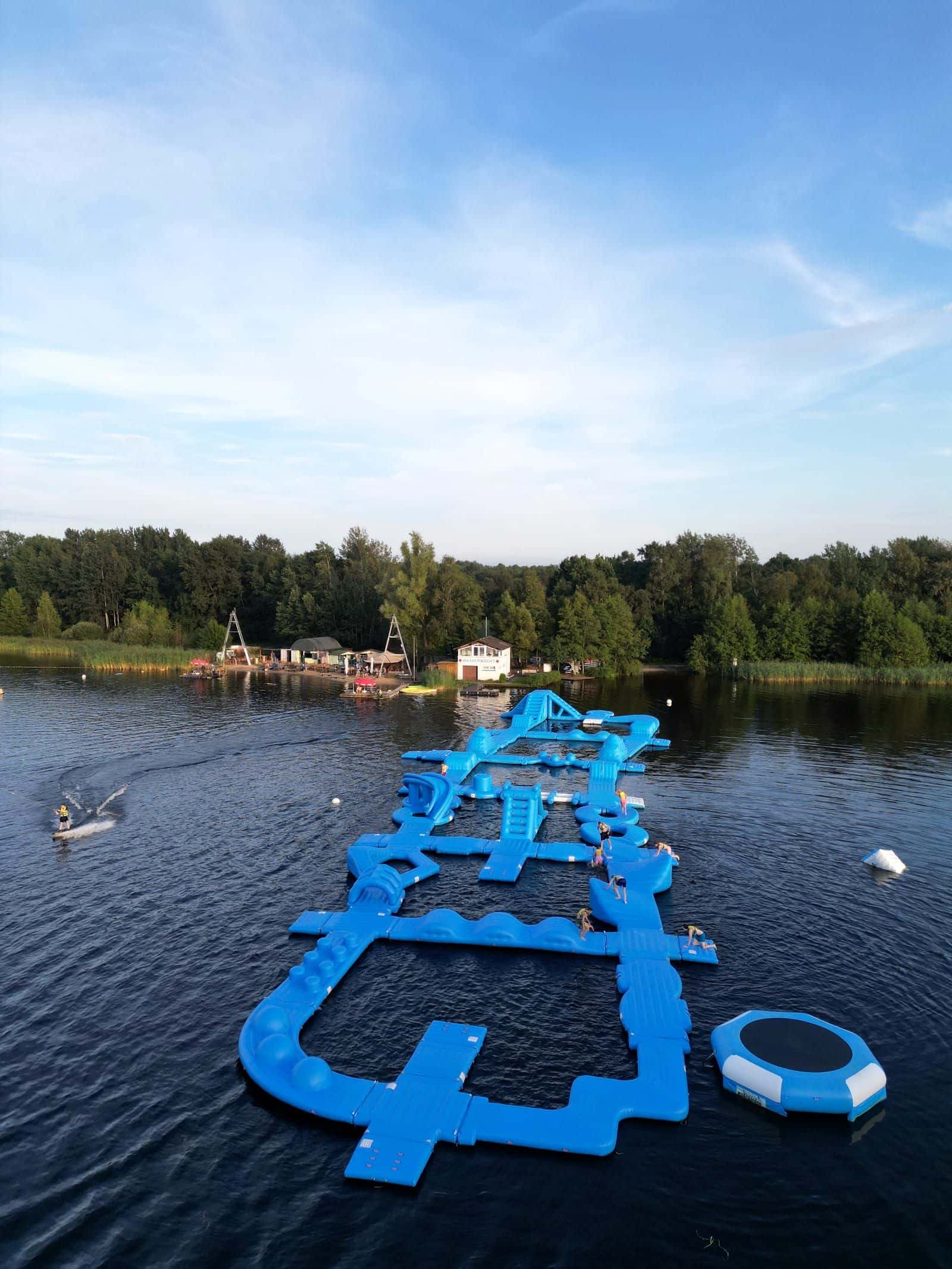 Luftaufnahme im Hochformat: der blaue Aquapark auf dem Oyter See, am Ufer Strand und Beachclub, links ein Wakeboarder