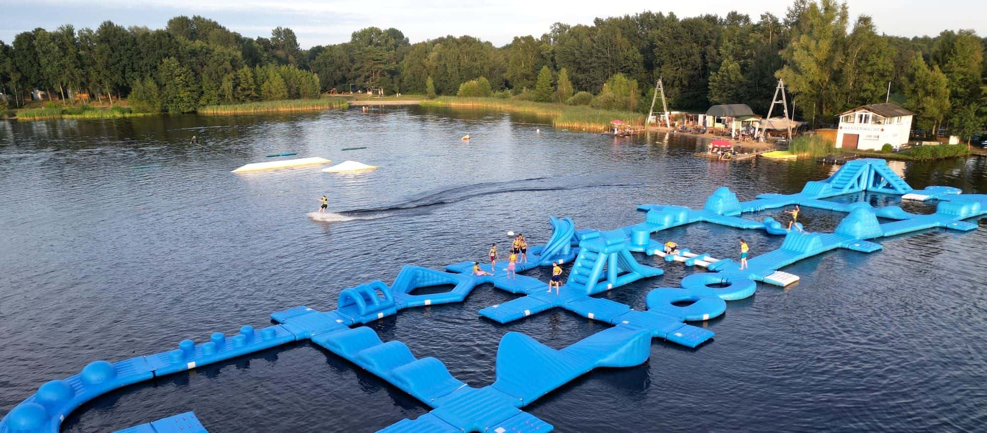 Weiter Blick über den Oyter See: Aquapark mit Gästen darauf, ein Wakeboarder am Seil, am Ufer Strand und Gebäude