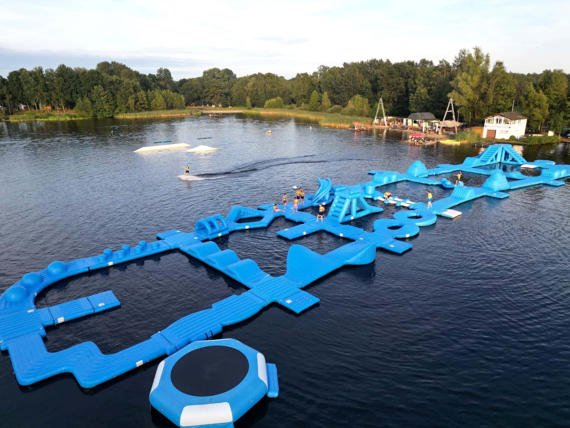 Luftaufnahme des Oyter Sees: blauer Aquapark mit Rutsche und Trampolin, mehrere Gäste auf dem Parcours, links ein Wakeboarder am Seil