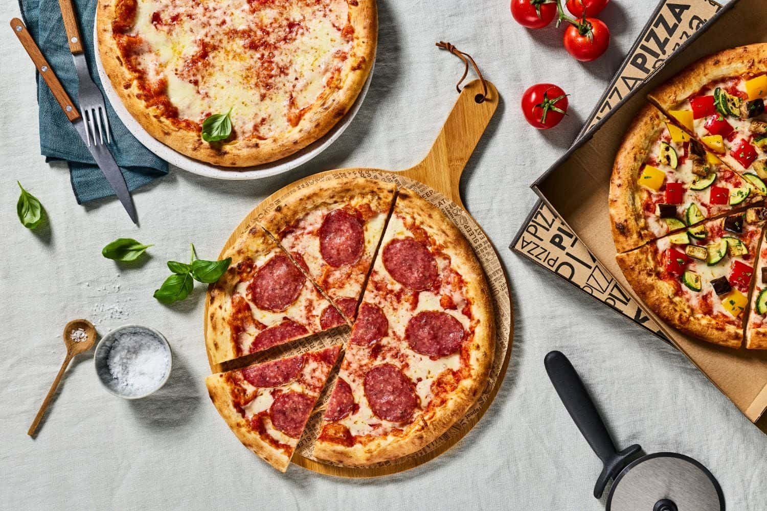 Von oben: Salami-Pizza in Stücken auf einem Holzbrett, daneben eine Margherita und eine Gemüsepizza im Karton