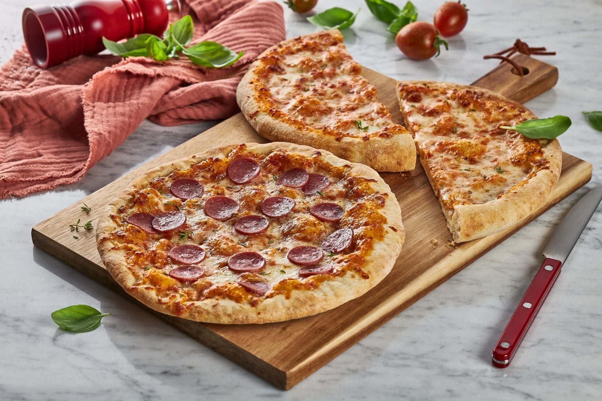Salami-Pizza und eine halbierte Margherita auf einem Holzbrett, daneben Basilikum, Kirschtomaten und ein Messer