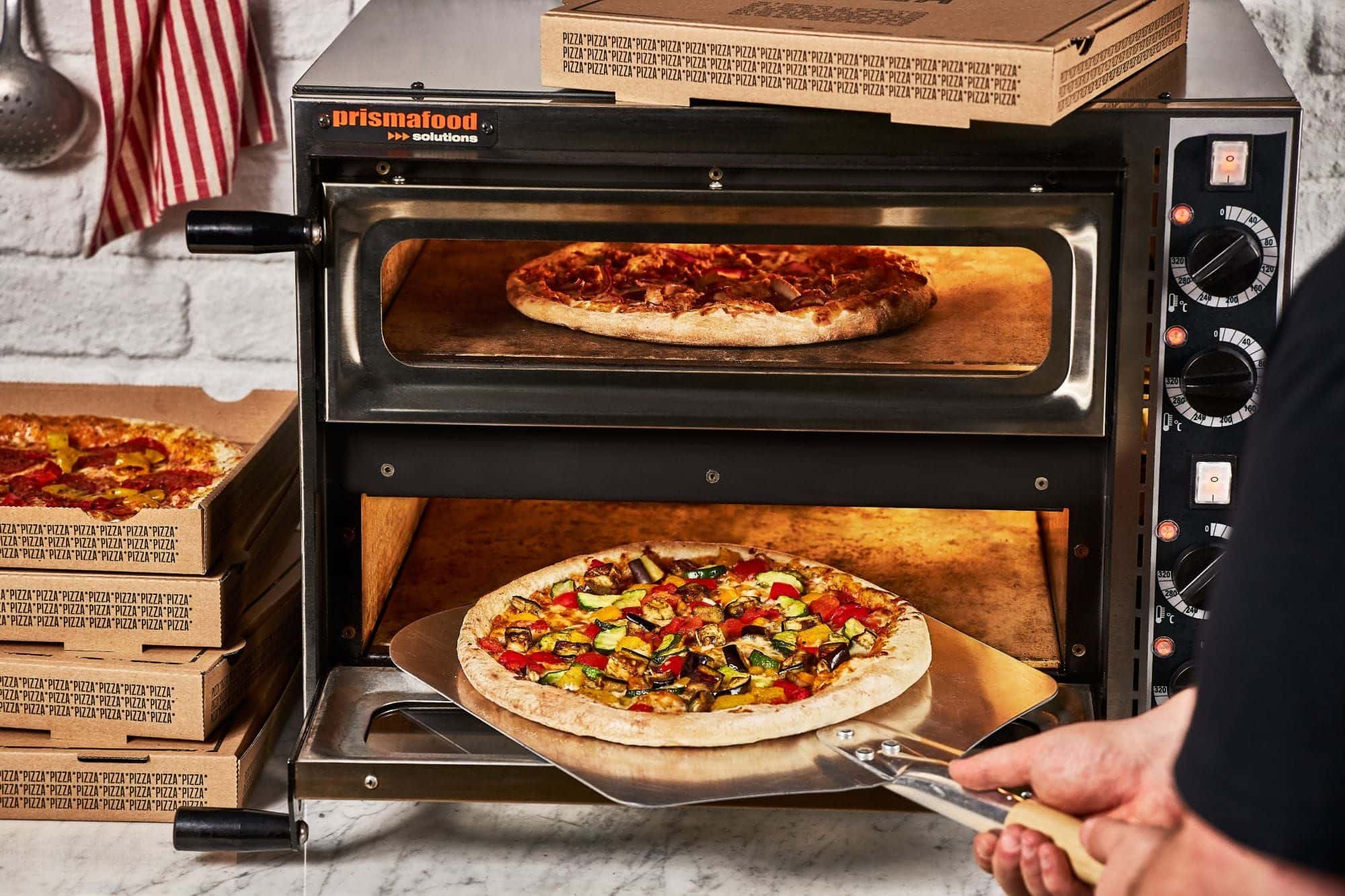 Eine Gemüsepizza wird mit dem Pizzaschieber in den Ofen geschoben, im oberen Fach backt eine zweite Pizza, daneben gestapelte Pizzakartons