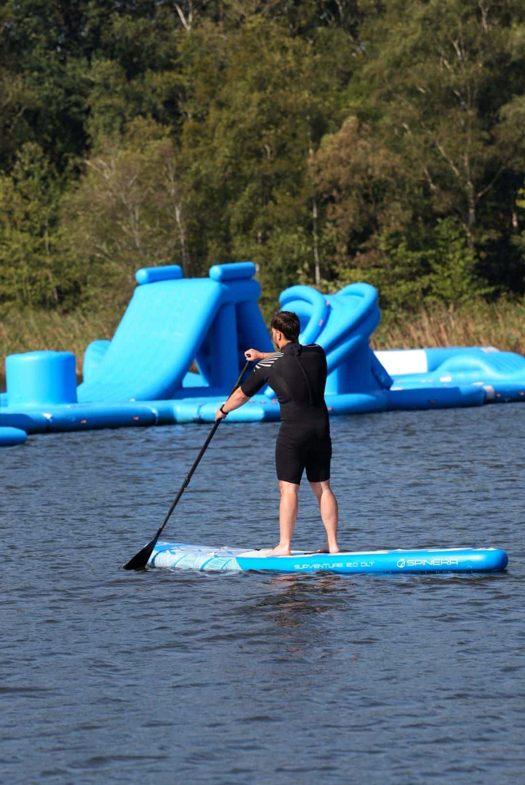 Mann von hinten im schwarzen Neopren auf einem blauen SUP-Board, vor ihm die Rutsche des Aquaparks