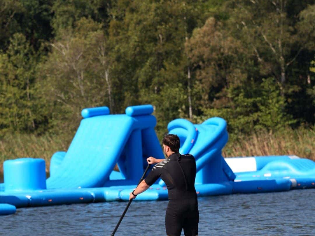 Mann im Neoprenanzug paddelt auf einem SUP-Board vor der blauen Rutsche des Aquaparks