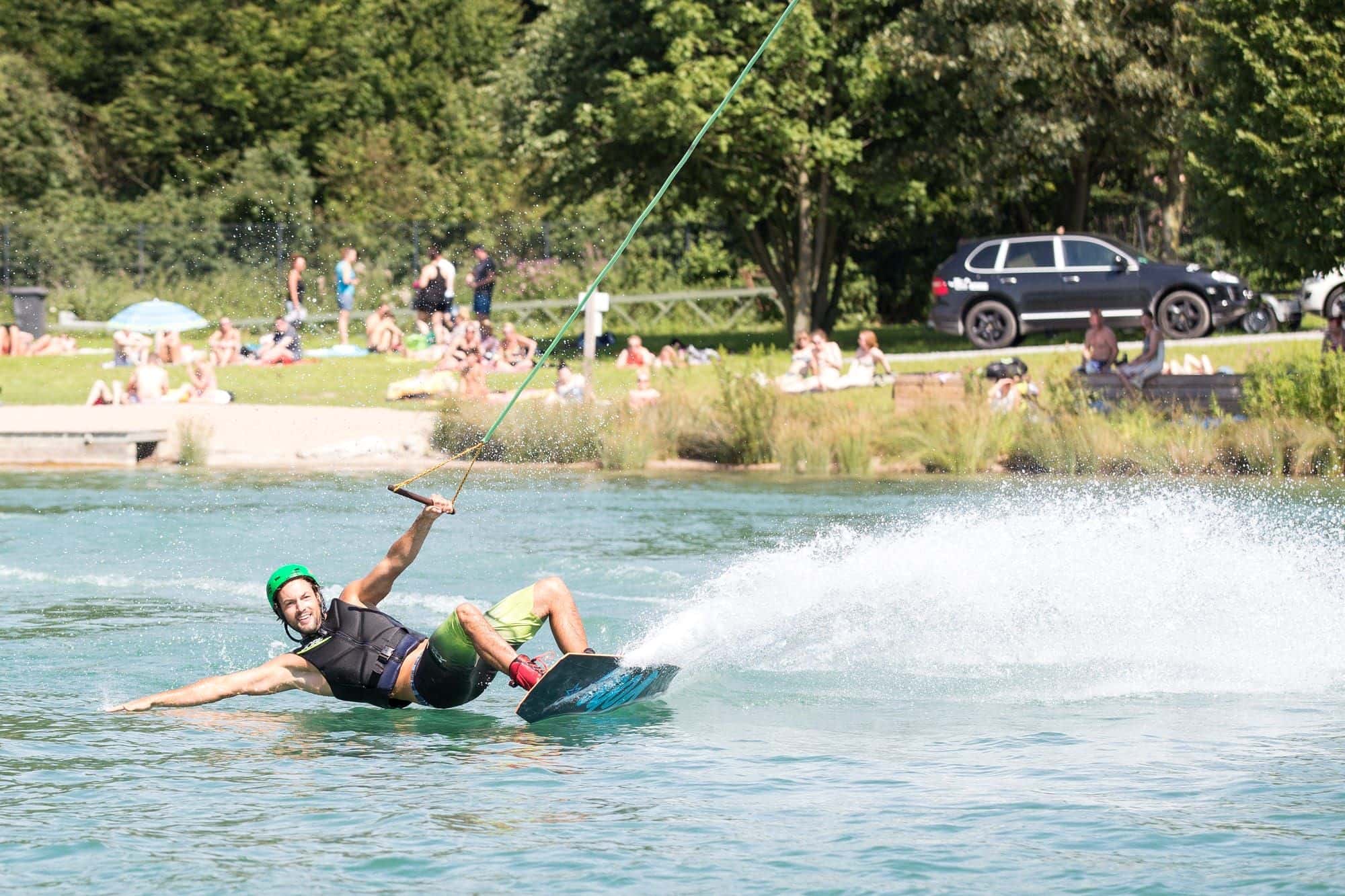Mann mit grünem Helm und schwarzer Weste legt sich in die Kurve, das Wakeboard wirft eine Gischtwand auf