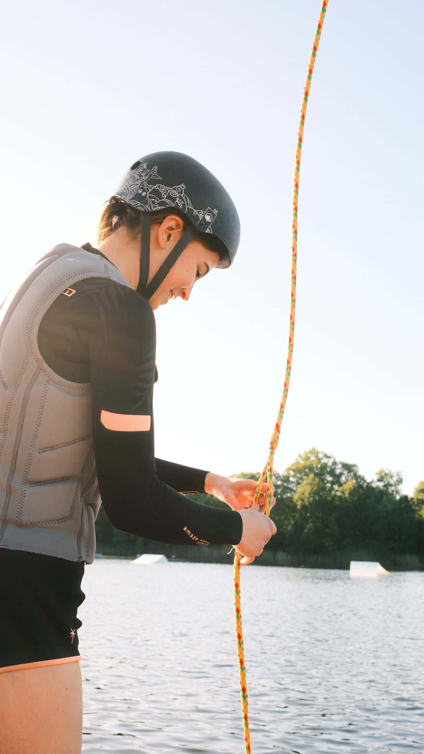 Junge Frau mit Helm und grauer Weste fädelt das bunte Seil am Steg ein, im Hintergrund der See