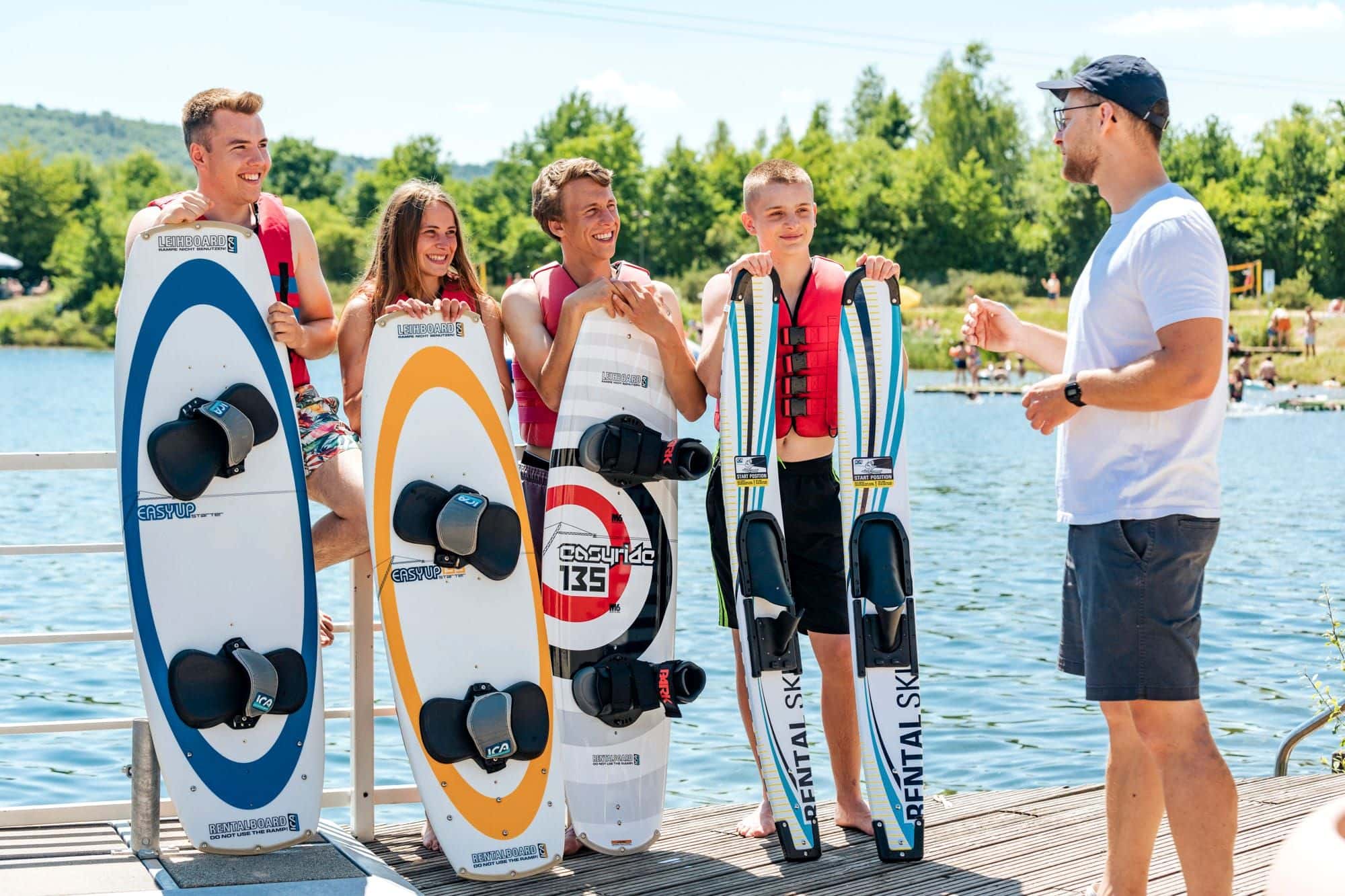 Vier Jugendliche mit Wakeboards und Wasserskiern hören am Steg einem Trainer im weißen Shirt zu