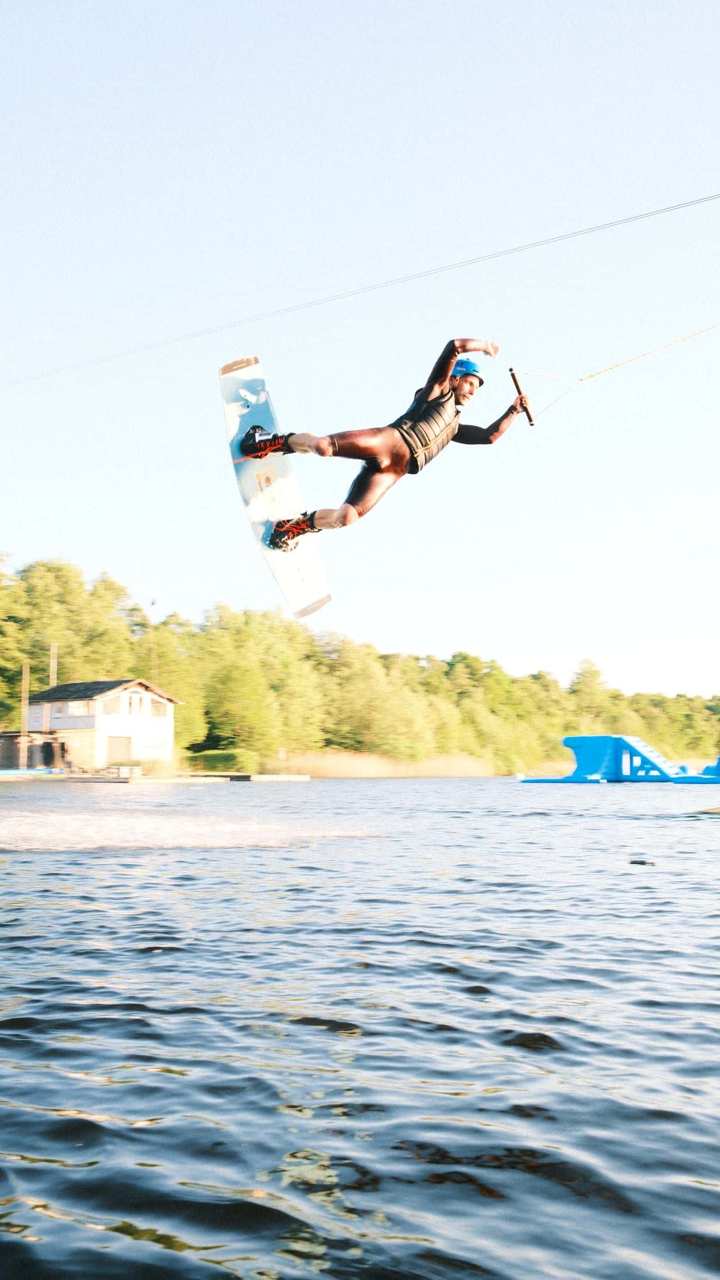 Wakeboarder im schwarzen Neopren und blauem Helm in der Luft bei einem Trick an der Seilbahn über dem Oyter See