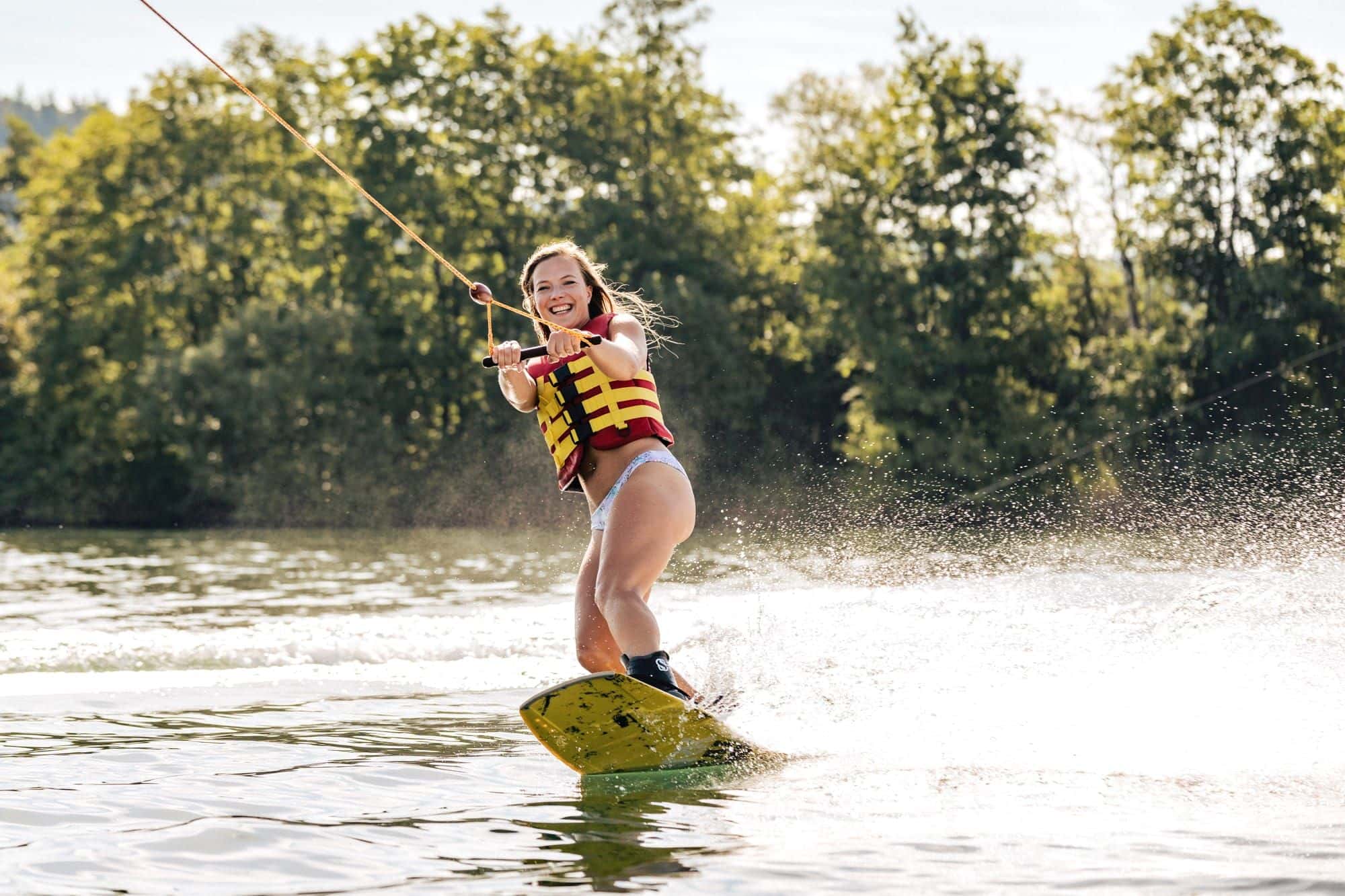 Lächelndes Mädchen mit gelb-roter Schwimmweste fährt Wakeboard, das gelbe Board wirft Gischt auf