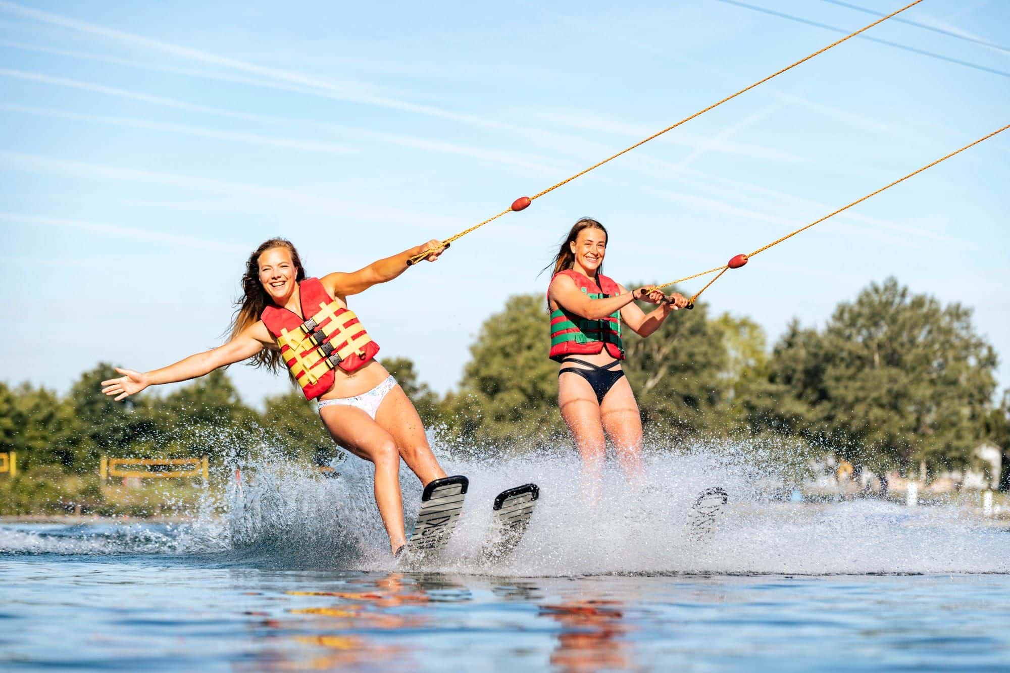 Zwei junge Frauen mit roten Schwimmwesten fahren nebeneinander Wasserski und lachen in die Gischt