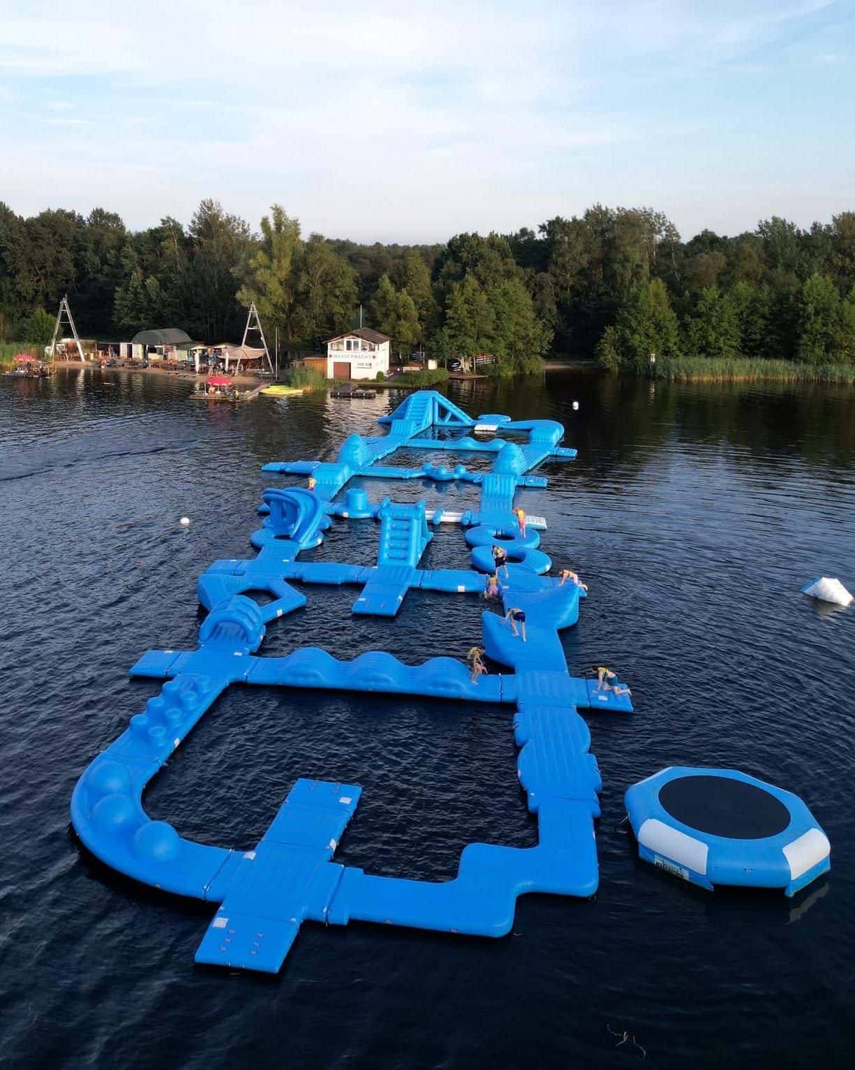Luftaufnahme im Hochformat: der blaue Aquapark mit Gästen auf dem Oyter See, dahinter Strand, Beachclub und Waldufer