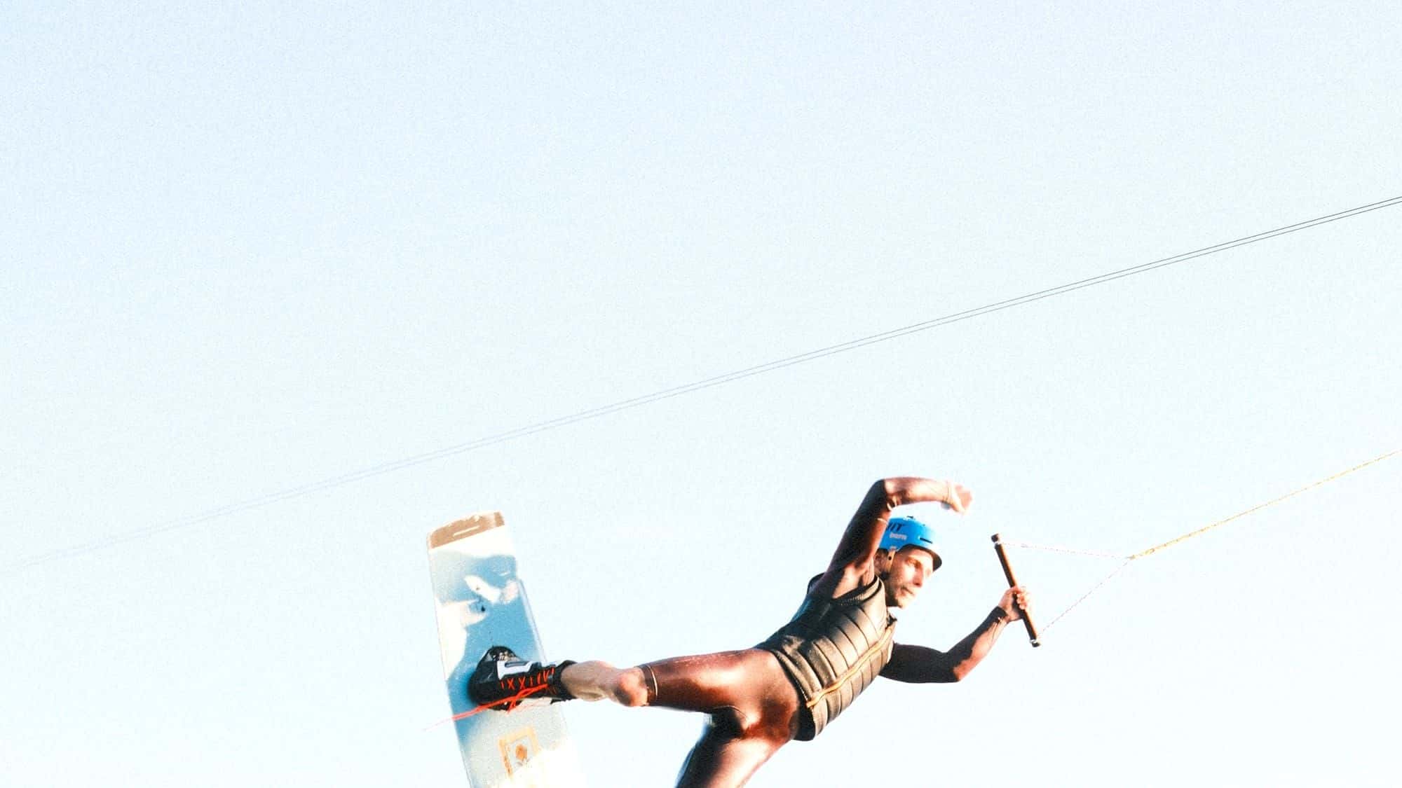 Wakeboarder im schwarzen Neopren und blauem Helm springt mit dem Board hoch in die Luft, dahinter heller Himmel und das Seil der Bahn